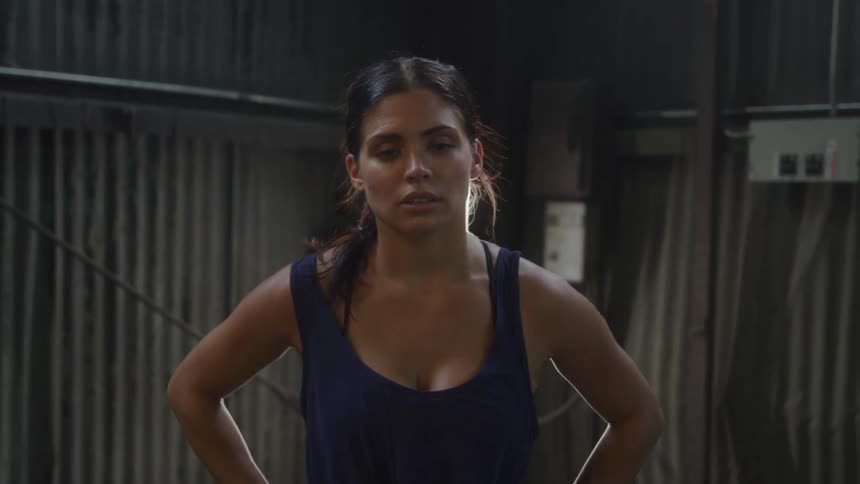 A woman with her dark hair pulled back stands in dimly lith warehouse wearing a navy blue tank top looking directly at the camera with her hands on her hips
