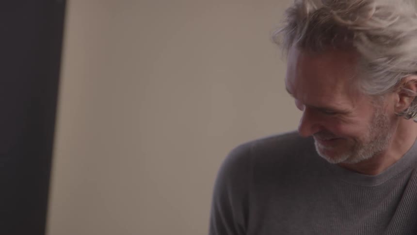 A smiling man with graying hair is looking at someone just offcamera he is wearing a gray longsleeved shirt and appears to be laughing or enjoying a joke