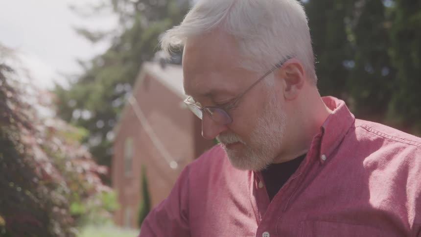 A middleaged man with graying hair and glasses is shown outdoors looking down he is wearing a pinkishred collared shirt the background shows a house and greenery on a sunny day