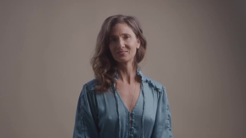 Woman with wavy brown hair wears a blue blouse and smiles slightly against a neutral background
