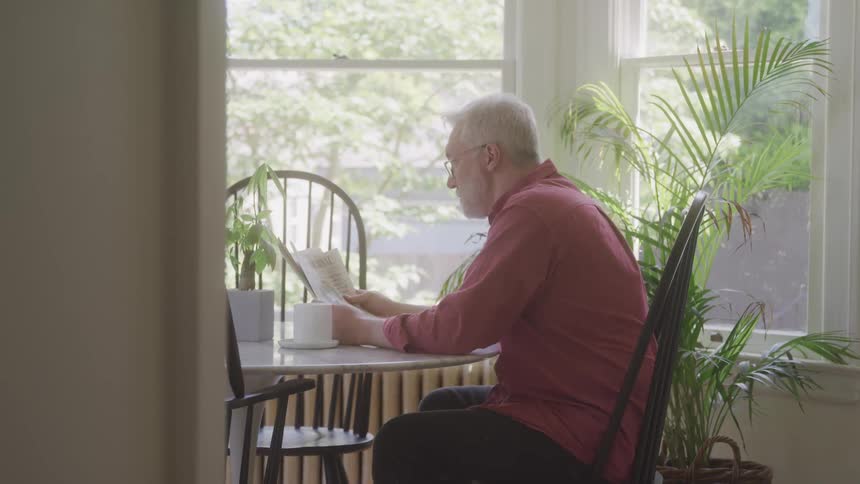 An older man in a red shirt sits at a table by the window reading a newspaper surrounded by houseplants
