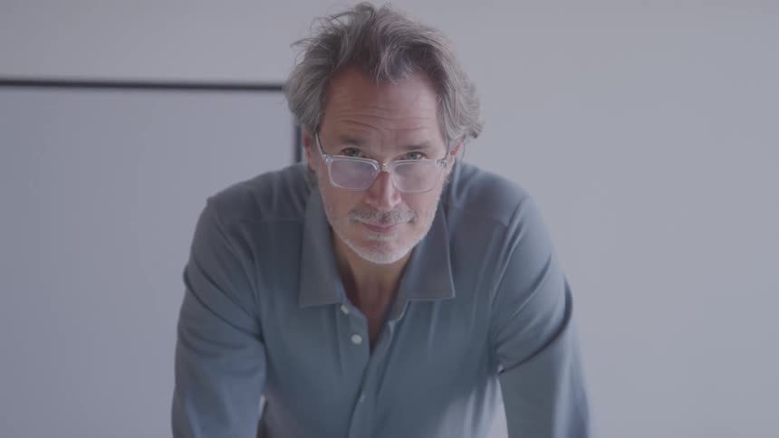 A man with grey hair and glasses looks down and then up at the camera with a serious expression