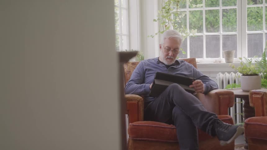 A man with a white beard and gray hair is sitting in an orange armchair looking at a tablet he is in a bright room with a large window and several potted plants