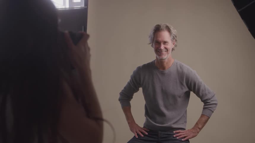 A man smiles confidently with hands on hips as a photographer captures his portrait in a studio