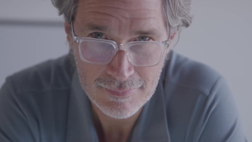 A man with a grey beard and glasses looks down and then up at the camera with a smile