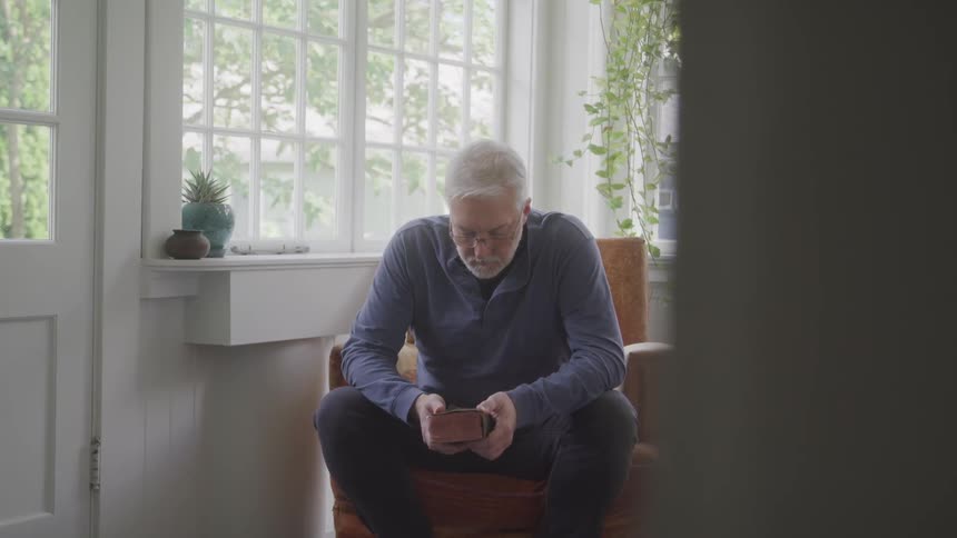 A cinematic shot from a partially obscured perspective shows an elderly man sitting in a sunlit room holding a book