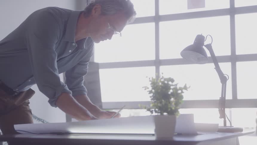 Man reviews project blueprints leaning on a desk lit by natural light