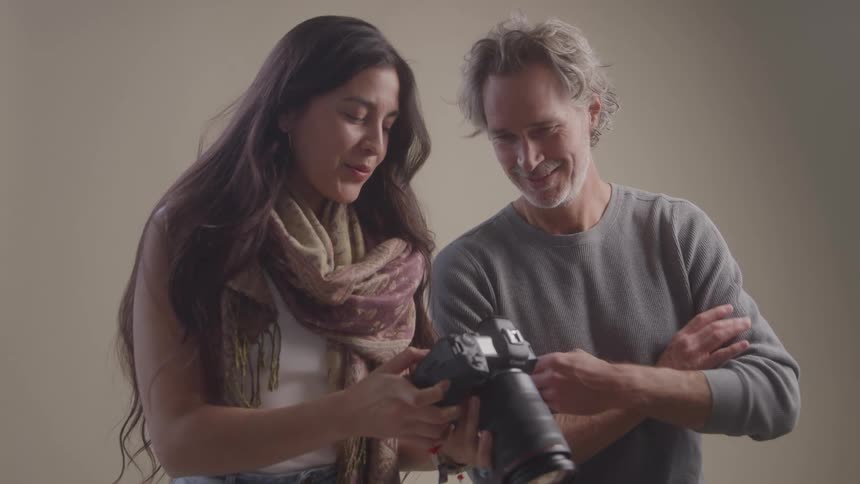 A woman and a man are looking at a camera together the woman is pointing at something on the cameras screen while the man smiles and watches