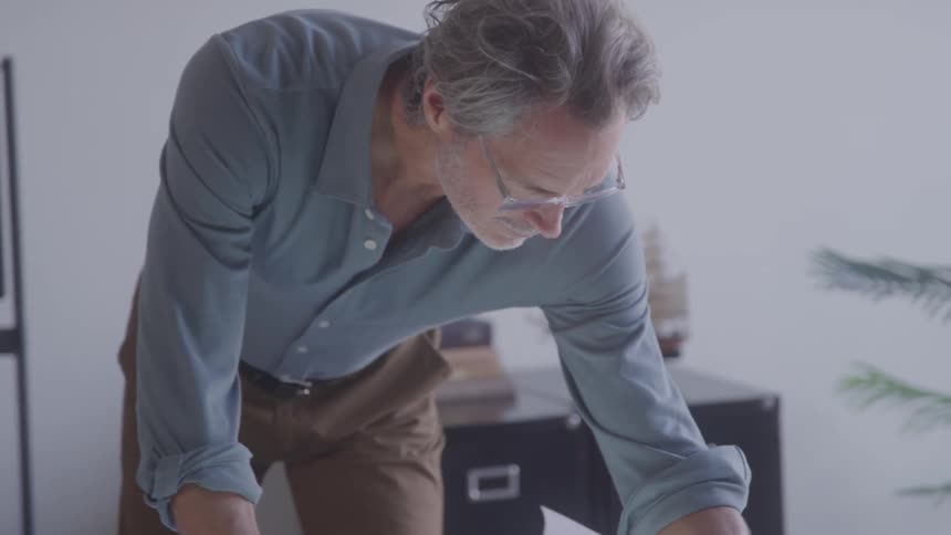 An older man standing in an office leaning over to read a sheet on a desk