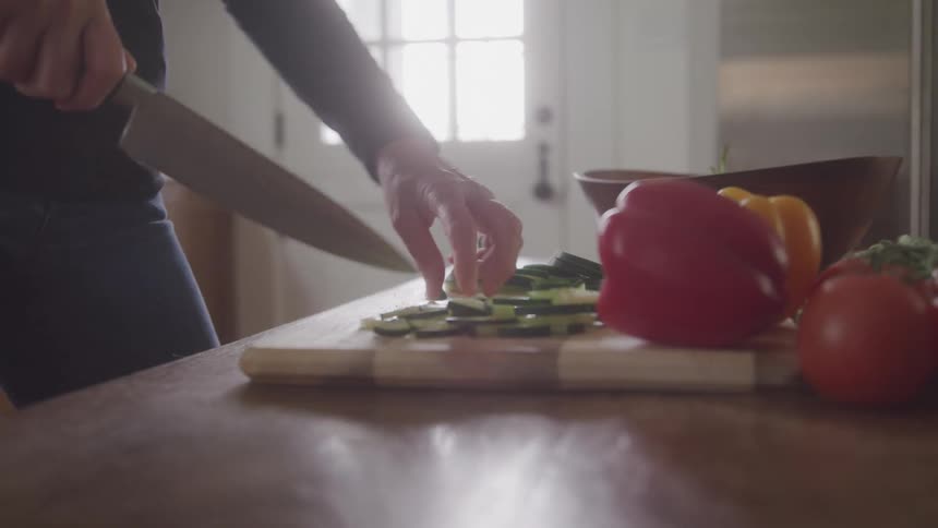 A person holds a large knife and slices a green vegetable on a wooden cutting board with a red pepper a yellow pepper and a tomato sitting next to it