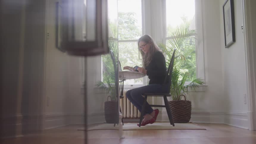 A woman with long blonde hair and glasses sits in a wooden chair at a small table working on a laptop with her feet resting on a basket