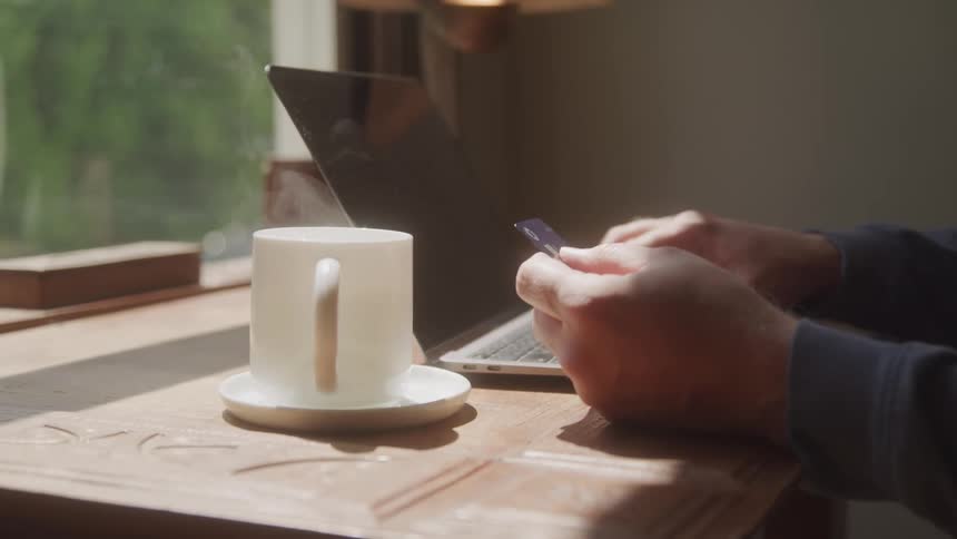 Pair of hands interacting with a laptop and holding a credit card on top of a table with a white cup