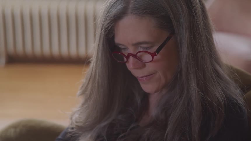 A woman with long gray hair and red glasses looks down with a focused expression in a softly lit room