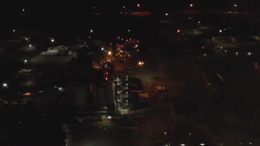An aerial view at night shows a large industrial complex with buildings and bright lights there is a plume of smoke or steam rising from the center of the complex