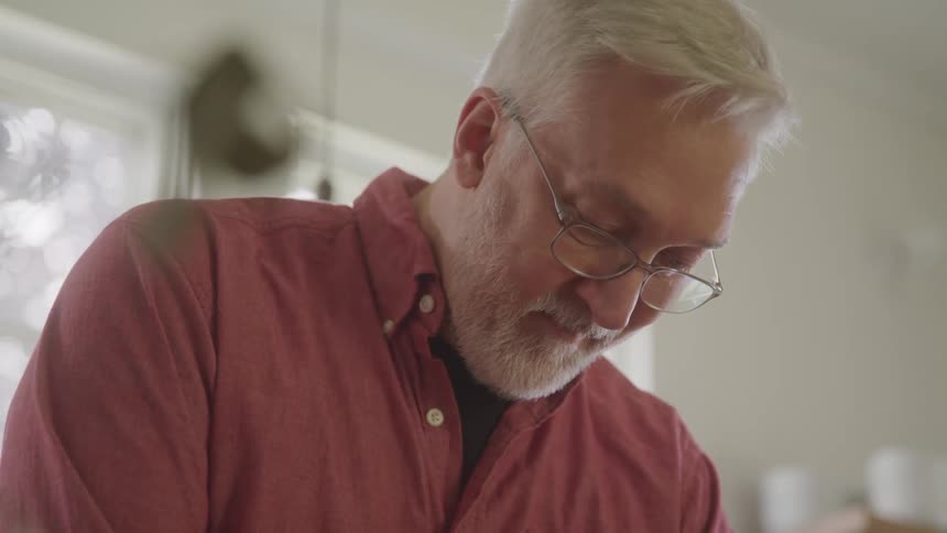 Greyhaired man wearing glasses and a red shirt focuses on an activity in a welllit indoor setting