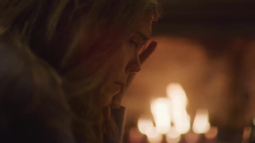 A pensive woman sits alone in a chair bathed in the warm flickering light of a nearby fireplace her hand resting near her face in a thoughtful pose