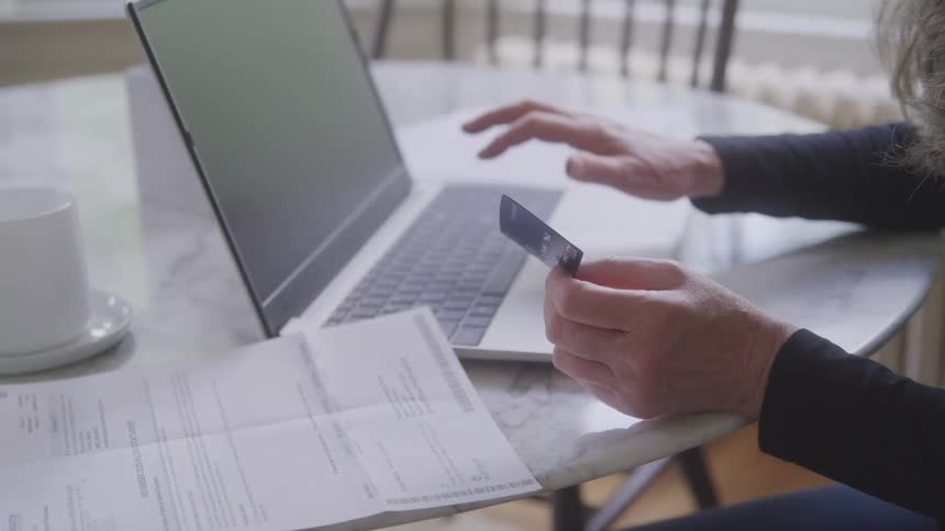 The video shows a closeup of a persons hands holding a credit card and using a laptop the person is sitting at a table with papers and a coffee mug next to them