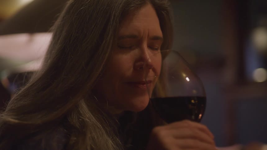 The video shows a closeup of a womans face with her eyes closed as she holds a glass of red wine she appears to be savoring the aroma and is in a restaurant or dimly lit bar