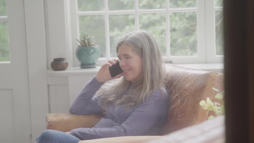The video shows an elderly woman with gray hair sitting in a chair talking on a flip phone she is positioned in a sunlit room with a window and potted plants visible behind her