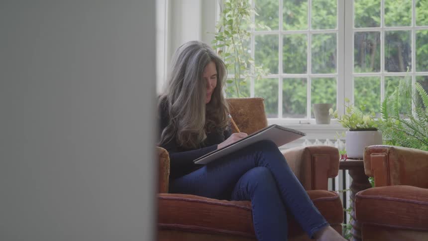 A female is sitting in a chair in a sun room writing on paper