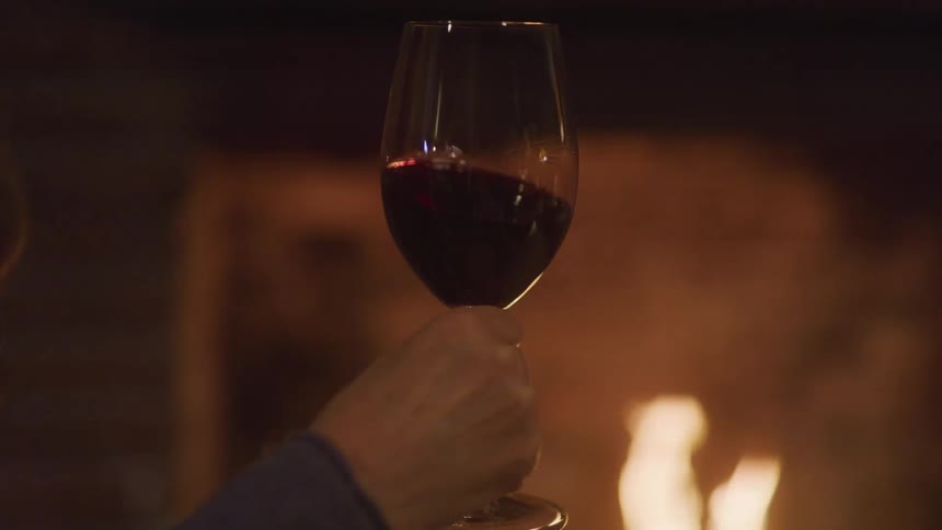 Glass of red wine raised in front of a lit fireplace