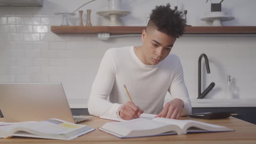 A black man is writing on paper with a book in front and a laptop at his side