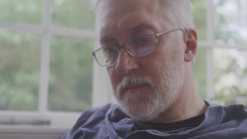A man with white hair and glasses looks down in quiet focus near a window with natural light