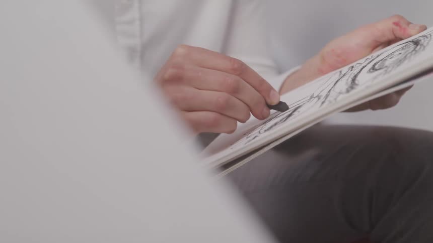 A person wearing a long sleeved white shirt and a watch is drawing a sketch in a notebook with a stick of charcoal
