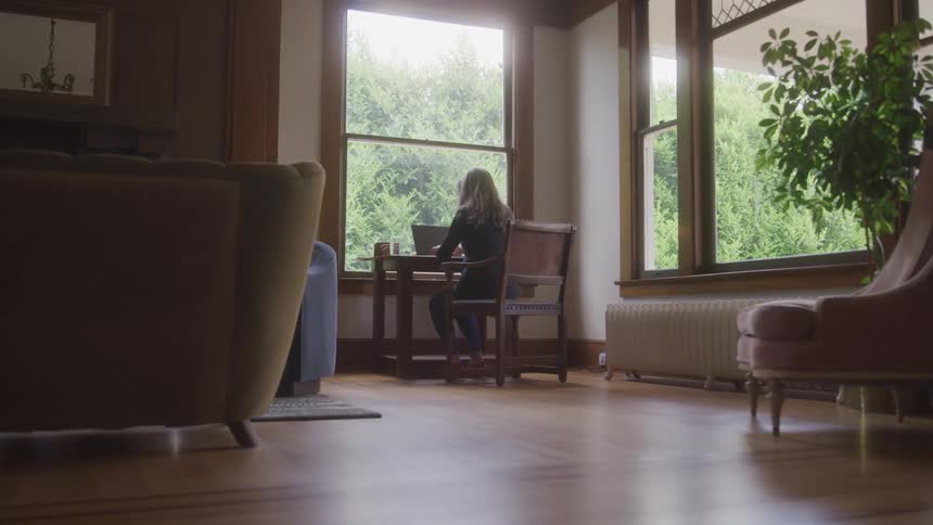Woman works on a laptop while sitting at a table near a large window overlooking trees