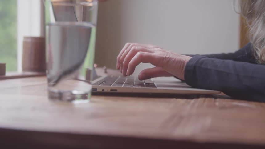 A person types on a laptop at a wooden table near a window