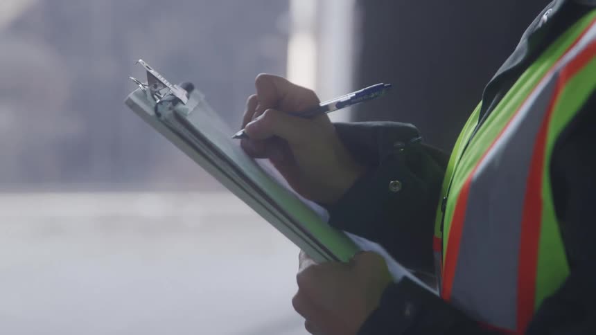 A closeup shot of a person in a highvisibility vest writing on a clipboard with a pen focused on the task