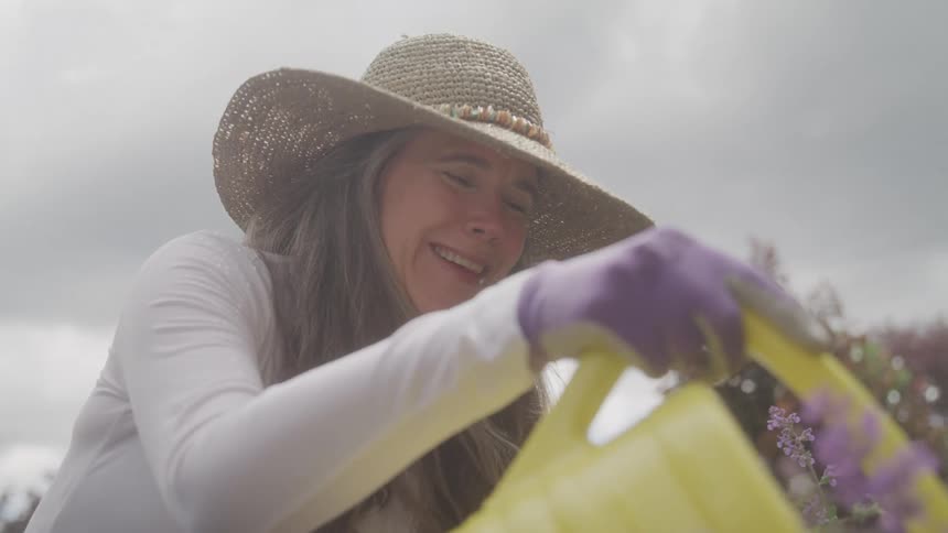 Woman wearing a straw hat smiles while watering flowers with a yellow watering can