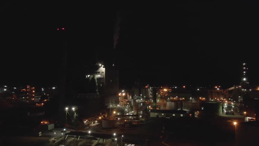 An aerial shot shows an industrial plant at night brightly lit against the dark sky smoke or steam is rising from one of the tall structures