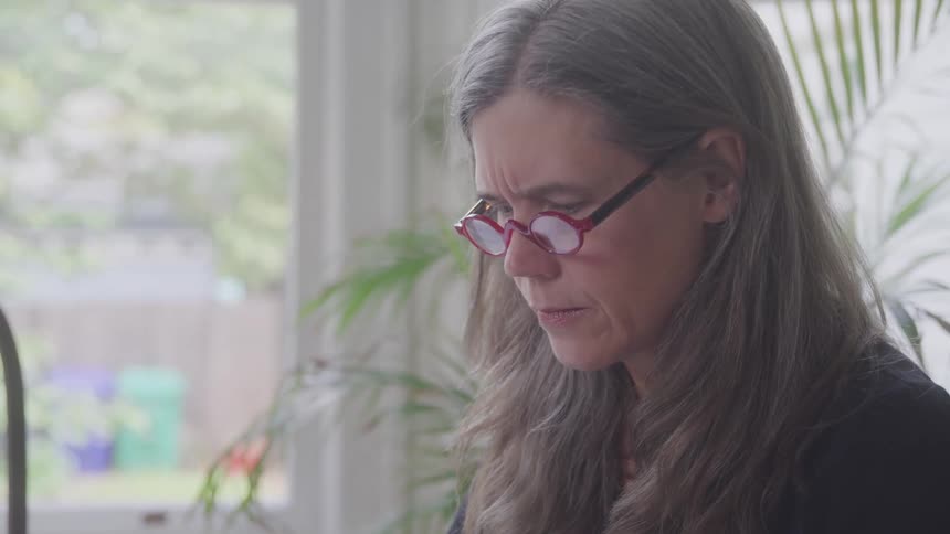 A woman with long grey hair and red framed glasses looks down at a paper and a window and a plant are visible in the background
