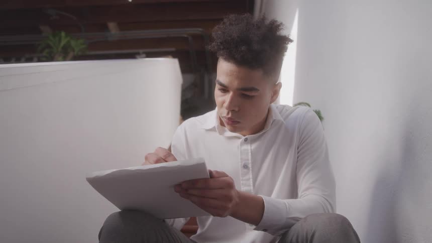 A teenage boy with short curly hair and a white shirt sits on a white staircase looking intently at a stack of papers he holds in his hands