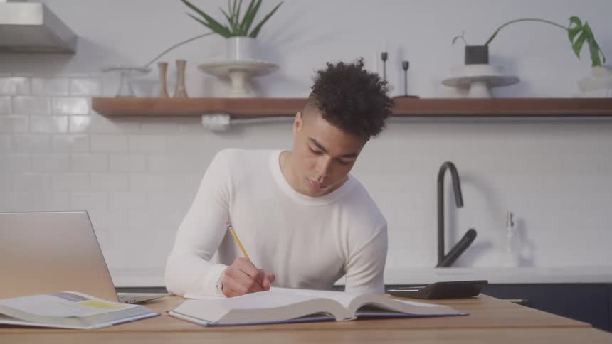 A student with short hair in a white long sleeved shirt sits at a wooden table doing homework
