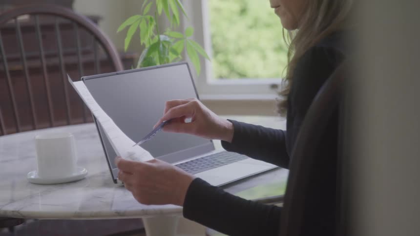 A woman sits at a table holding a receipt and credit card beside an open laptop