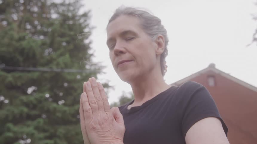 A tranquil medium shot of a person with their eyes closed and hands in a prayer position meditating or doing yoga in an outdoor suburban setting