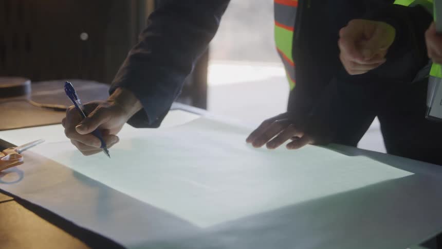 Hand holds a blue pen while drawing on a lighted table with a technical plan