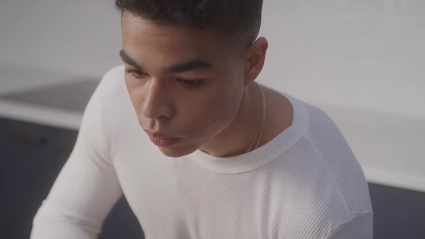 A young man with short curly hair is shown from the chest up looking down and to his right he is wearing a white ribbed longsleeve shirt and appears focused on something outside the frame