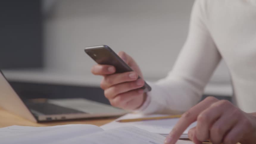 A person uses a smartphone while reviewing documents on a desk