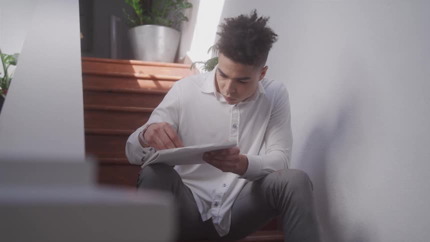 A young man with short curly hair and a white collared shirt sits on a set of stairs writing on a notebook
