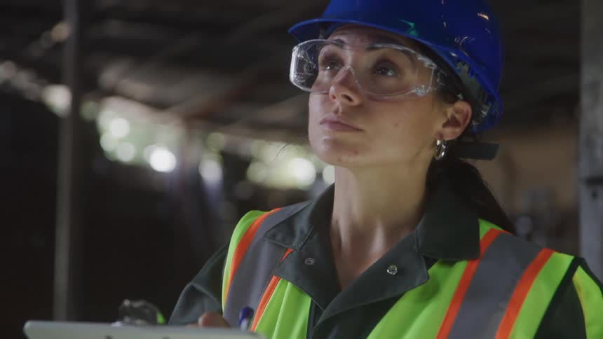 Woman wearing a safety vest blue helmet and holding a clipboard conducts an inspection in an industrial setting