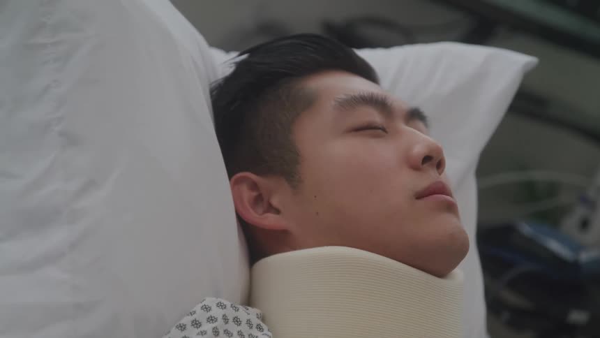 A closeup profile shot of an injured man wearing a neck brace with his eyes closed in a hospital bed