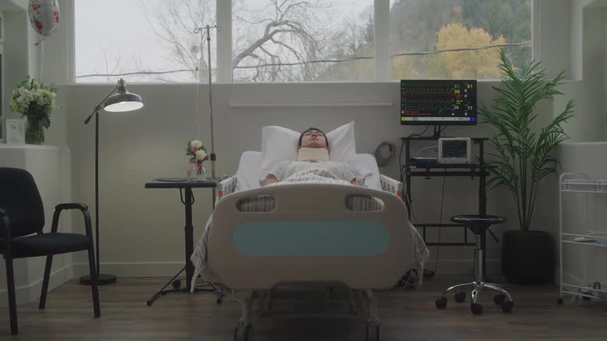 The video shows a patient lying in a hospital bed in a bright modern room the patient is seen resting under a blanket with medical equipment visible around them and a large window in the background shows a view of trees with yellow and green leaves