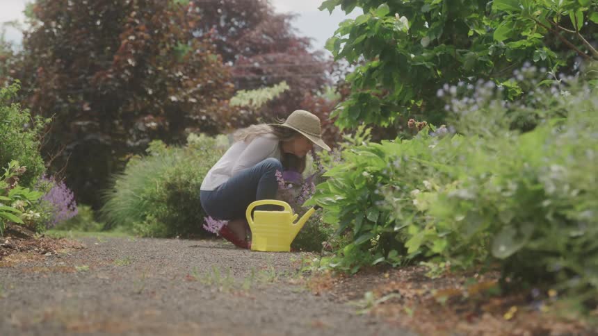 A woman in a sun hat crouches in a garden path tending to flowers beside a yellow watering can