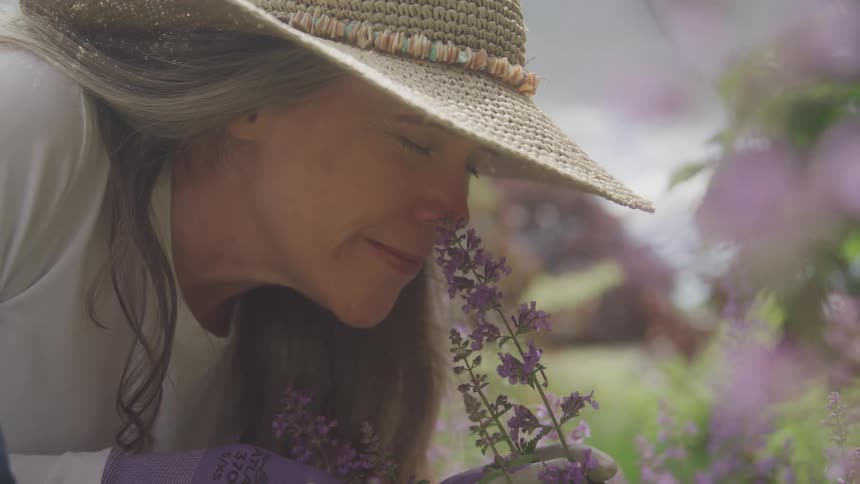 A woman in a widebrimmed hat leans close to purple flowers in a sunlit garden appearing to smell them