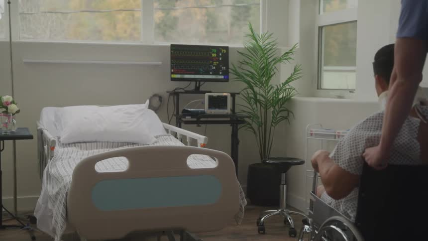 A medical professional pushes a patient wearing a neck brace in a wheelchair across a hospital room towards an empty bed