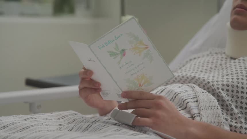 A female wearing a neck brace in a hospital bed opens a greeting card to read