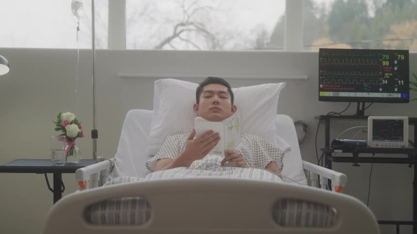 A young asian male patient reads a card while lying in bed in a hospital during daytime